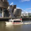 Un barco turístico se desliza a lo largo del río cerca de un edificio moderno y un imponente rascacielos en Bilbao, ofreciendo la experiencia perfecta de Paseo en barco por Bilbao en un día despejado. - ForTwo - Planes para parejas
