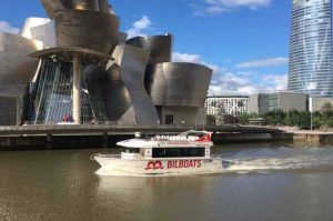Un barco turístico se desliza a lo largo del río cerca de un edificio moderno y un imponente rascacielos en Bilbao, ofreciendo la experiencia perfecta de Paseo en barco por Bilbao en un día despejado. - ForTwo - Planes para parejas