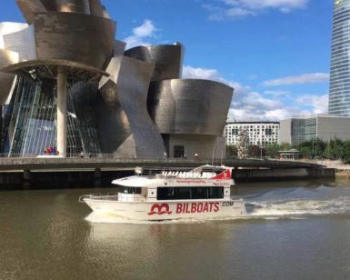 Un barco turístico se desliza a lo largo del río cerca de un edificio moderno y un imponente rascacielos en Bilbao, ofreciendo la experiencia perfecta de Paseo en barco por Bilbao en un día despejado. - ForTwo - Planes para parejas