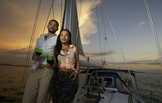 Una pareja disfruta de un romántico paseo en barco en la Herradura y Almuñécar, tomando bebidas mientras se deslizan por aguas serenas con velas puestas contra la vibrante puesta de sol. - ForTwo - Planes para parejas