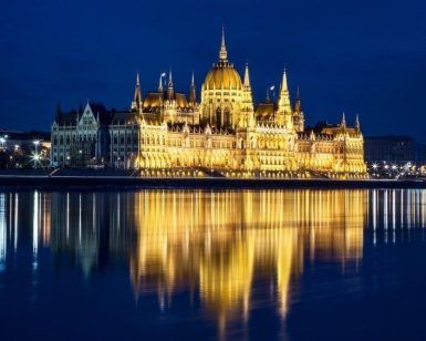 El edificio junto al río iluminado por la noche crea reflejos románticos en el agua, creando una escena pintoresca perfecta para el "Romántico paseo en barco al anochecer (Budapest)". - ForTwo - Planes para parejas