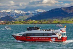 Un barco turístico ofrece un encantador "Paseo en barco en Santander" en un lago sereno, con montañas nevadas y exuberantes colinas verdes que evocan la belleza de Santander en el fondo. - ForTwo - Planes para parejas