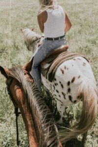 Una mujer con una camiseta blanca sin mangas cabalga sobre un caballo moteado por los exuberantes campos de hierba cerca de Córdoba, con el encanto histórico de Medina Azahara como telón de fondo en la "Ruta a caballo por la zona de Medina Azahara (Córdoba)". Se ve parcialmente otro caballo. - ForTwo - Planes para parejas