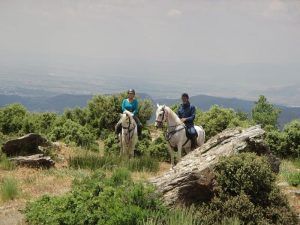 Dos personas disfrutan de un paseo a caballo por la ladera de una colina en Güéjar Sierra, donde las amplias vistas del valle y las lejanas montañas de Sierra Nevada crean el escenario perfecto para una inolvidable Escapada rural hípica de 2 noches a Sierra Nevada en Güéjar Sierra. - ForTwo - Planes para parejas