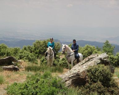 Dos personas disfrutan de un paseo a caballo por la ladera de una colina en Güéjar Sierra, donde las amplias vistas del valle y las lejanas montañas de Sierra Nevada crean el escenario perfecto para una inolvidable Escapada rural hípica de 2 noches a Sierra Nevada en Güéjar Sierra. - ForTwo - Planes para parejas