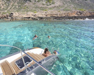La gente nada cerca de un yate en las cristalinas aguas turquesas de una costa rocosa, ideal para "Sorprende a tu pareja con un paseo en catamarán" en Palma de Mallorca. - ForTwo - Planes para parejas