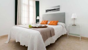 Un dormitorio minimalista exhibe una cama grande con cabecera gris y almohadas naranjas, combinada con una mesa auxiliar adornada con una lámpara y una bandeja decorativa, creando un ambiente tan tranquilo como una "Escapada con desayuno y crucero en barco por el Guadalquivir (Sevilla). - ForTwo - Planes para parejas