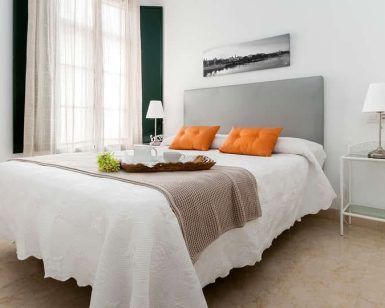 Un dormitorio minimalista exhibe una cama grande con cabecera gris y almohadas naranjas, combinada con una mesa auxiliar adornada con una lámpara y una bandeja decorativa, creando un ambiente tan tranquilo como una "Escapada con desayuno y crucero en barco por el Guadalquivir (Sevilla). - ForTwo - Planes para parejas