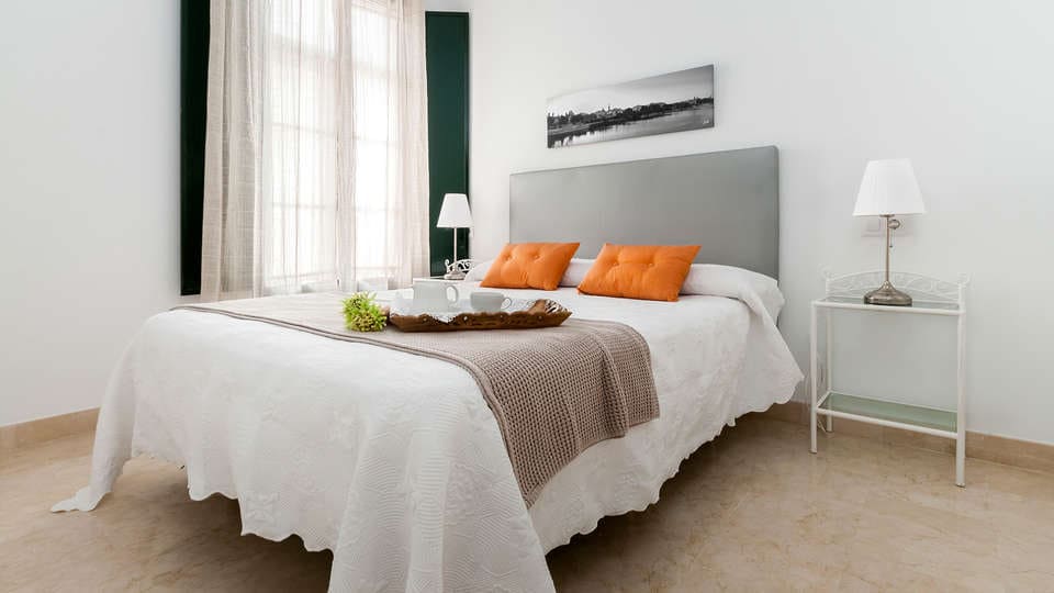 Un dormitorio minimalista exhibe una cama grande con cabecera gris y almohadas naranjas, combinada con una mesa auxiliar adornada con una lámpara y una bandeja decorativa, creando un ambiente tan tranquilo como una "Escapada con desayuno y crucero en barco por el Guadalquivir (Sevilla). - ForTwo - Planes para parejas