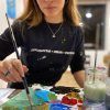 Una persona con una camisa negra y un texto creativo pinta utilizando una paleta vibrante, encarnando la esencia viva de "Clase de pintura con tu copita de vino en Arte Bar (Barcelona). - ForTwo - Planes para parejas