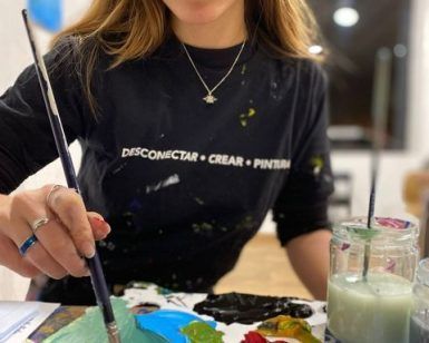 Una persona con una camisa negra y un texto creativo pinta utilizando una paleta vibrante, encarnando la esencia viva de "Clase de pintura con tu copita de vino en Arte Bar (Barcelona). - ForTwo - Planes para parejas