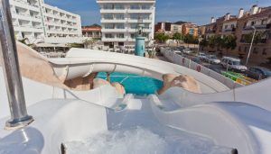 Desde la cima de un emocionante paseo en el Planazo de toboganes y almuerzo buffet en Pineda Splash 30º Hotels en Barcelona, se abre la vibrante escena; una piscina brillante debajo contrasta con edificios distantes y autos estacionados. - ForTwo - Planes para parejas