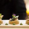 En La Bodeguita de María de Valencia, el chef presenta artísticamente un plato de pescado gourmet adornado con verdes coloridos y flores violetas en un plato blanco impecable. - ForTwo - Planes para parejas