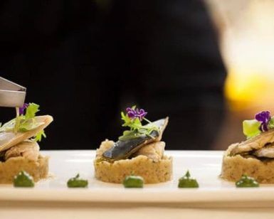 En La Bodeguita de María de Valencia, el chef presenta artísticamente un plato de pescado gourmet adornado con verdes coloridos y flores violetas en un plato blanco impecable. - ForTwo - Planes para parejas