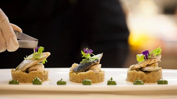 En La Bodeguita de María de Valencia, el chef presenta artísticamente un plato de pescado gourmet adornado con verdes coloridos y flores violetas en un plato blanco impecable. - ForTwo - Planes para parejas