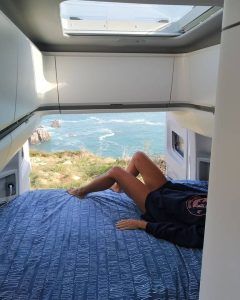 Relajándose en un acogedor Alquiler de furgonetas camper con Mundovan (Badajoz), la persona disfruta de una vista impresionante a través de la puerta trasera abierta, donde la costa rocosa se encuentra con el océano infinito. - ForTwo - Planes para parejas