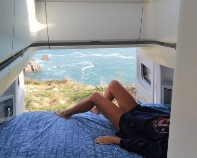 Relajándose en un acogedor Alquiler de furgonetas camper con Mundovan (Badajoz), la persona disfruta de una vista impresionante a través de la puerta trasera abierta, donde la costa rocosa se encuentra con el océano infinito. - ForTwo - Planes para parejas