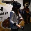 Una persona con gafas de realidad virtual y blazer, inmersa en la Experiencia de videojuegos en realidad virtual con PlayVR (Barcelona), sostiene unos mandos frente a una pared decorada. - ForTwo - Planes para parejas