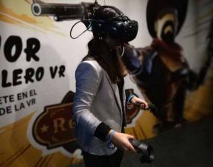 Una persona con gafas de realidad virtual y blazer, inmersa en la Experiencia de videojuegos en realidad virtual con PlayVR (Barcelona), sostiene unos mandos frente a una pared decorada. - ForTwo - Planes para parejas