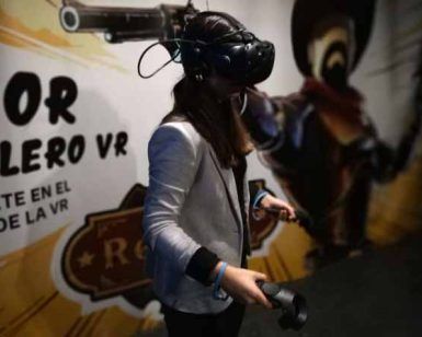 Una persona con gafas de realidad virtual y blazer, inmersa en la Experiencia de videojuegos en realidad virtual con PlayVR (Barcelona), sostiene unos mandos frente a una pared decorada. - ForTwo - Planes para parejas