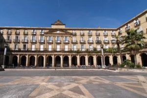 Un gran edificio con arcos y columnas, enmarcado por balcones y palmeras bajo un cielo azul vibrante, invita a la exploración a través del "Free tour por Bilbao. - ForTwo - Planes para parejas