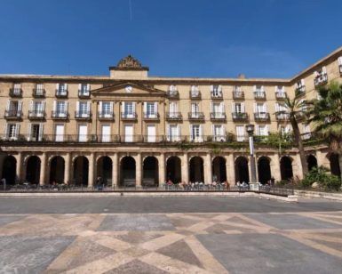 Un gran edificio con arcos y columnas, enmarcado por balcones y palmeras bajo un cielo azul vibrante, invita a la exploración a través del "Free tour por Bilbao. - ForTwo - Planes para parejas