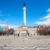 Experimente la majestuosidad de una alta columna blanca coronada por una estatua en una plaza decorada, abrazada por encantadores edificios y frondosos árboles, bajo el cielo azul de Lisboa con nuestro "Tour gratuito guiado en Lisboa". - ForTwo - Planes para parejas