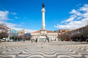 Experimente la majestuosidad de una alta columna blanca coronada por una estatua en una plaza decorada, abrazada por encantadores edificios y frondosos árboles, bajo el cielo azul de Lisboa con nuestro "Tour gratuito guiado en Lisboa". - ForTwo - Planes para parejas