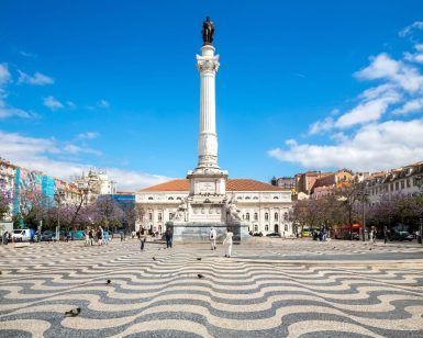 Experimente la majestuosidad de una alta columna blanca coronada por una estatua en una plaza decorada, abrazada por encantadores edificios y frondosos árboles, bajo el cielo azul de Lisboa con nuestro "Tour gratuito guiado en Lisboa". - ForTwo - Planes para parejas
