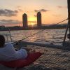 Relajándose en la red de un catamarán, la persona disfruta de la Puesta del sol en Barcelona desde Catamarán o velero, contemplando el cálido resplandor del atardecer sobre el horizonte de Barcelona con dos altos edificios al fondo. - ForTwo - Planes para parejas