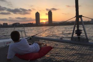 Relajándose en la red de un catamarán, la persona disfruta de la Puesta del sol en Barcelona desde Catamarán o velero, contemplando el cálido resplandor del atardecer sobre el horizonte de Barcelona con dos altos edificios al fondo. - ForTwo - Planes para parejas