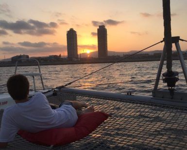 Relajándose en la red de un catamarán, la persona disfruta de la Puesta del sol en Barcelona desde Catamarán o velero, contemplando el cálido resplandor del atardecer sobre el horizonte de Barcelona con dos altos edificios al fondo. - ForTwo - Planes para parejas
