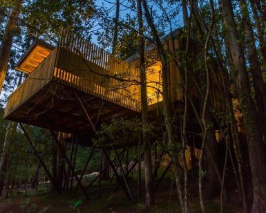 El Punta Espiñeiro, el lujo de Cabanas sem Barreiras en A Coruña, ofrece un escape sereno con una casa en el árbol elevada sobre pilotes entre los árboles, que brilla cálidamente a través de grandes ventanales al anochecer, proporcionando una tranquilidad similar a las acogedoras cabañas. - ForTwo - Planes para parejas