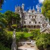 Explore una mansión histórica ornamentada con una fachada detallada, exuberante vegetación y estatuas en el recorrido Visita a Sintra y Cascais con entrada a Quinta da Regaleira, frente a un cielo azul vibrante. - ForTwo - Planes para parejas