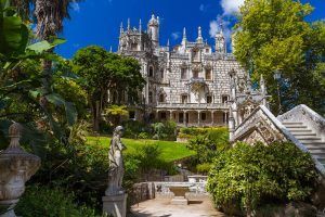Explore una mansión histórica ornamentada con una fachada detallada, exuberante vegetación y estatuas en el recorrido Visita a Sintra y Cascais con entrada a Quinta da Regaleira, frente a un cielo azul vibrante. - ForTwo - Planes para parejas
