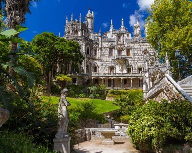 Explore una mansión histórica ornamentada con una fachada detallada, exuberante vegetación y estatuas en el recorrido Visita a Sintra y Cascais con entrada a Quinta da Regaleira, frente a un cielo azul vibrante. - ForTwo - Planes para parejas