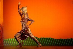 Un artista disfrazado de león captura la esencia del *Musical El Rey León en Madrid* en el escenario, con un fondo naranja y una escenografía verde vibrante, dando vida a la magia de este querido espectáculo. - ForTwo - Planes para parejas