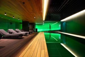 Descubra la "Sensación de serenidad" en el Spa Mandarin Oriental de Barcelona. Disfrute de una moderna piscina cubierta con iluminación ambiental verde, terraza de madera y tumbonas tejidas, dispuestas para capturar la esencia de un tranquilo retiro de spa en cada detalle. - ForTwo - Planes para parejas