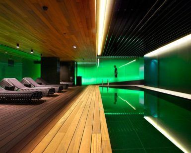Descubra la "Sensación de serenidad" en el Spa Mandarin Oriental de Barcelona. Disfrute de una moderna piscina cubierta con iluminación ambiental verde, terraza de madera y tumbonas tejidas, dispuestas para capturar la esencia de un tranquilo retiro de spa en cada detalle. - ForTwo - Planes para parejas