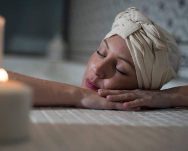 En Le Patio Spa, una persona disfruta de un Ritual Básico con exfoliación, con los ojos cerrados y la cabeza envuelta en una toalla, descansando sobre los brazos cruzados junto a velas encendidas para una relajación máxima. - ForTwo - Planes para parejas