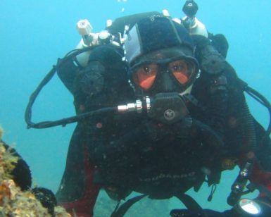 Un buceador, completamente equipado con equipo de buceo, disfruta de la emoción de la "Oferta: Bautismo de buceo (Granada)" mientras explora bajo el agua cerca de rocas cubiertas de coral y una vibrante vida marina. - ForTwo - Planes para parejas