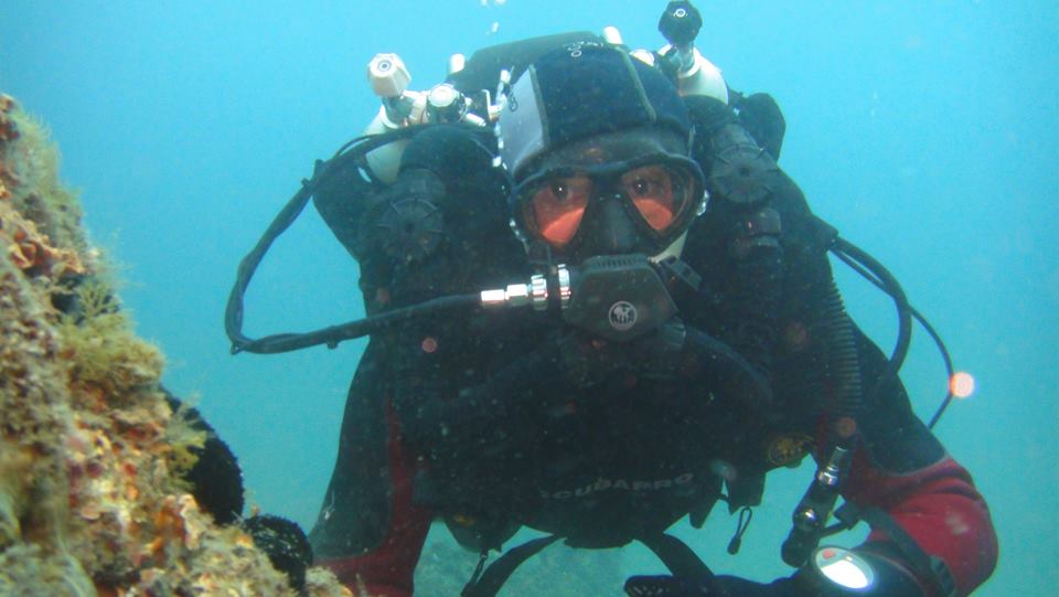 Un buceador, completamente equipado con equipo de buceo, disfruta de la emoción de la "Oferta: Bautismo de buceo (Granada)" mientras explora bajo el agua cerca de rocas cubiertas de coral y una vibrante vida marina. - ForTwo - Planes para parejas