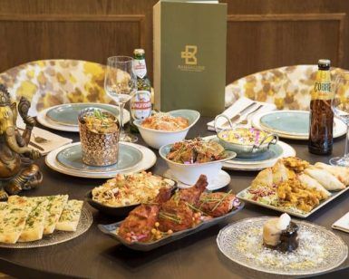 La mesa festiva de Sabores de India presenta la Oferta degustación Sabores de India: restaurante Bangalore (Madrid), completa con platos indios, botellas de cerveza y una estatua decorativa, que lleva el sabor de Bangalore al corazón de Madrid. - ForTwo - Planes para parejas