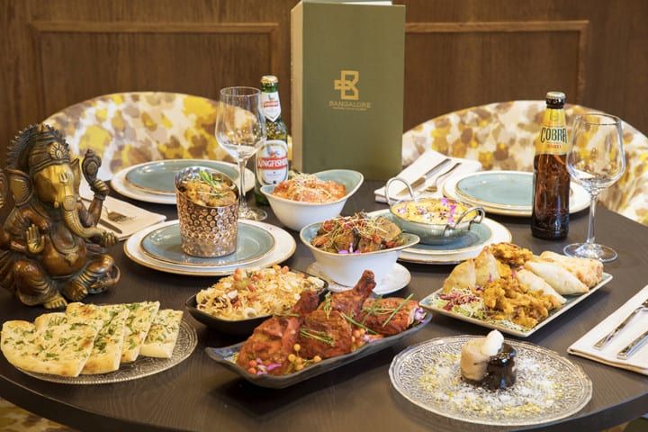 La mesa festiva de Sabores de India presenta la Oferta degustación Sabores de India: restaurante Bangalore (Madrid), completa con platos indios, botellas de cerveza y una estatua decorativa, que lleva el sabor de Bangalore al corazón de Madrid. - ForTwo - Planes para parejas