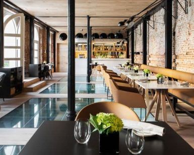 El Restaurante Dos Cielos en Madrid presume de un interior moderno con suelo de cristal, paredes de ladrillo y una elegante iluminación. Las mesas están elegantemente dispuestas con copas y plantas verdes, creando un ambiente refrescante. - ForTwo - Planes para parejas