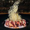 Presentado en un plato oscuro con una guarnición crujiente, este elegante plato de pulpo es perfecto para una cena romántica en La Tabernilla del Darro en Granada con un fondo rústico. - ForTwo - Planes para parejas