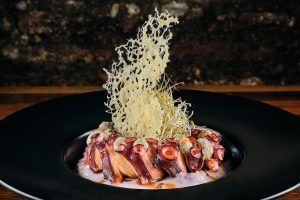 Presentado en un plato oscuro con una guarnición crujiente, este elegante plato de pulpo es perfecto para una cena romántica en La Tabernilla del Darro en Granada con un fondo rústico. - ForTwo - Planes para parejas