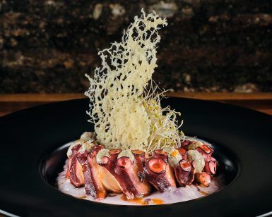 Presentado en un plato oscuro con una guarnición crujiente, este elegante plato de pulpo es perfecto para una cena romántica en La Tabernilla del Darro en Granada con un fondo rústico. - ForTwo - Planes para parejas