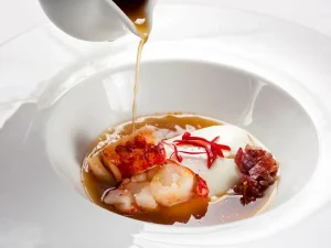 Disfrute de un menú degustación con estrella Michelin en el Restaurante Nectari de Barcelona, que incluye un plato gourmet de huevo escalfado, camarones y jamón en un caldo rico servido en un tazón blanco impecable. - ForTwo - Planes para parejas