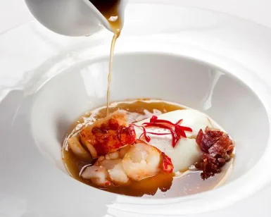 Disfrute de un menú degustación con estrella Michelin en el Restaurante Nectari de Barcelona, que incluye un plato gourmet de huevo escalfado, camarones y jamón en un caldo rico servido en un tazón blanco impecable. - ForTwo - Planes para parejas